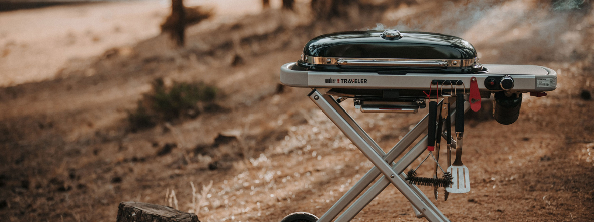 η φορητη ψησταριά υγραερίου Traveler σε ενα βουνό με διπλα εναν κορμό και γυρω γυρω φύλλα, απο την οποία βγαίνει καπνός και κρέμονται μια βούρτσα, μια λαβίδα και μια σπάτουλα